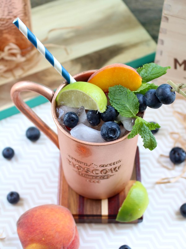 Copper mug containing Moscow Mule alcoholic drink, garnished with mint leaves, lime slices, peach slices, and blueberries