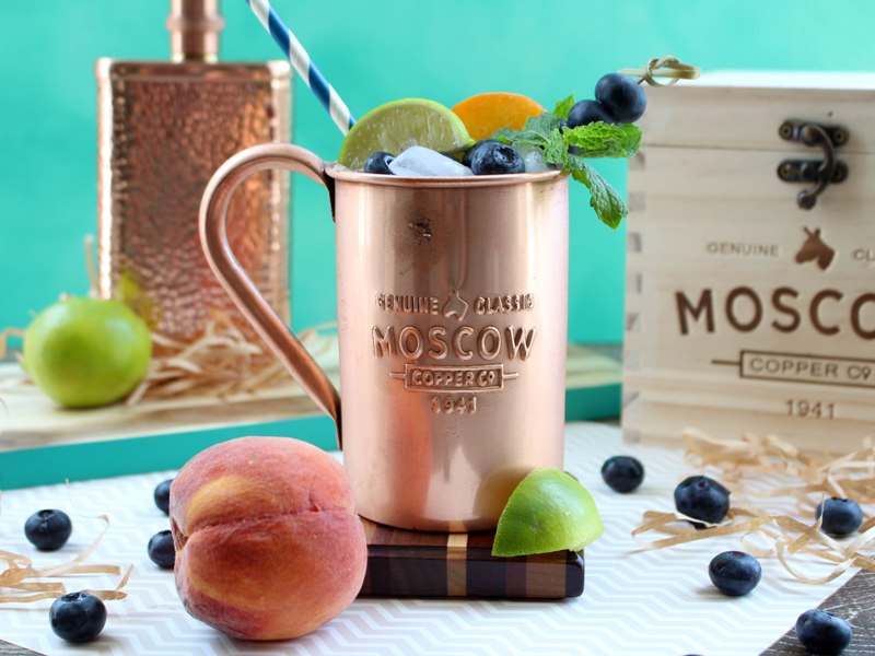 Copper mug containing Moscow Mule alcoholic drink, garnished with mint leaves, lime slices, peach slices, and blueberries. A wooden box and copper flask are in the background.