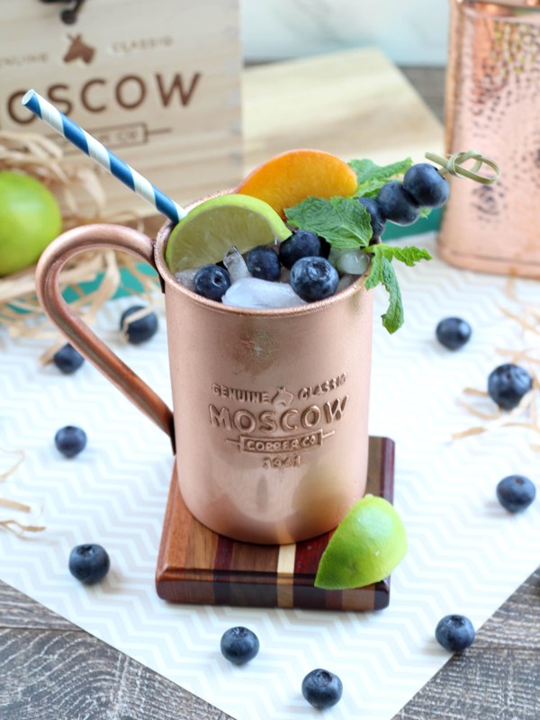 Cpper mug containing Moscow Mule alcoholic drink, garnished with mint leaves, lime slices, peach slices, and blueberries