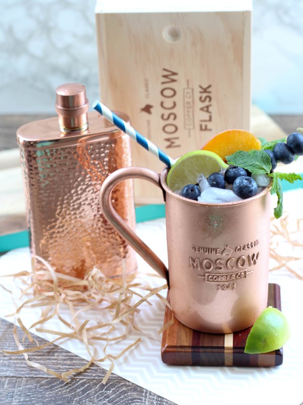 Copper mug containing Moscow Mule alcoholic drink, garnished with mint leaves, lime slices, peach slices, and blueberries. Next to mug is a hammered copper flask.