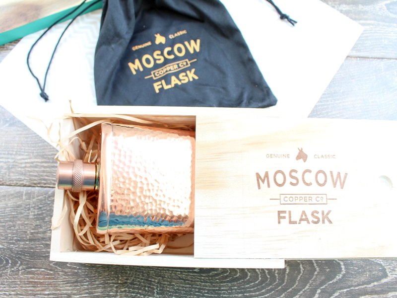 Wooden box containing hammered copper wooden flask, laying on top of a black velvet gift bag with "Moscow Flask" logo.