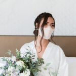Woman with dark hair wearing white face mask and white robe holding bouquet of white flowers