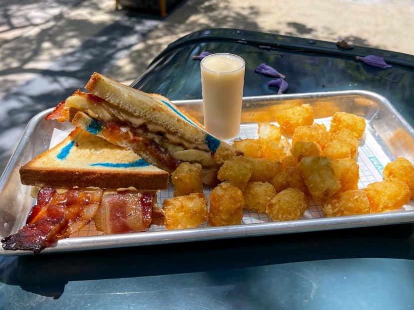 Sandwich with blue swirls in the bread and bacon hanging out next to side of crispy potato bites