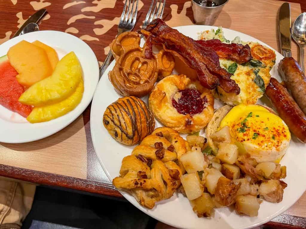 Two plates showing several food items from Storytellers Cafe brunch buffet, including fruit, Mickey waffle, bacon, potatoes, and danishes