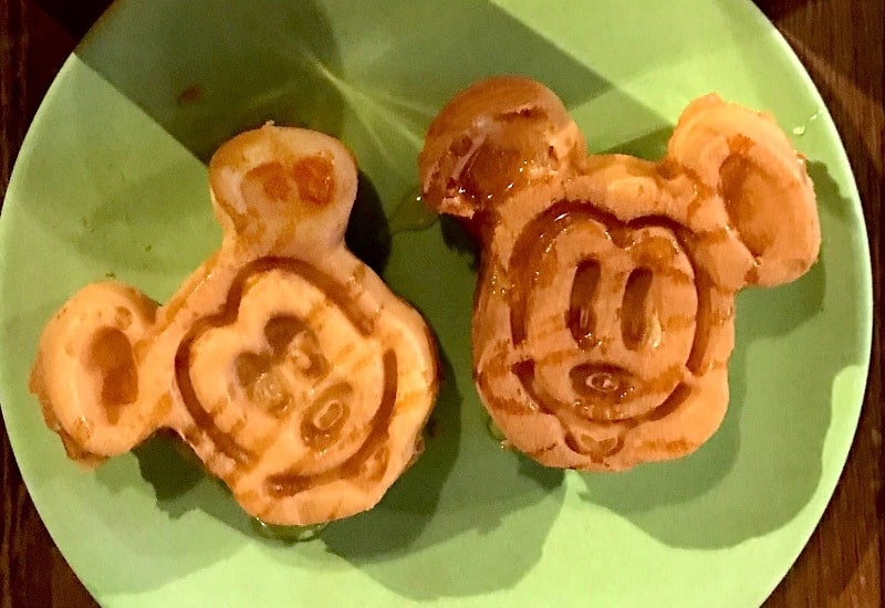 Two waffles shaped like Mickey Mouse drizzled with maple syrup on a green plate