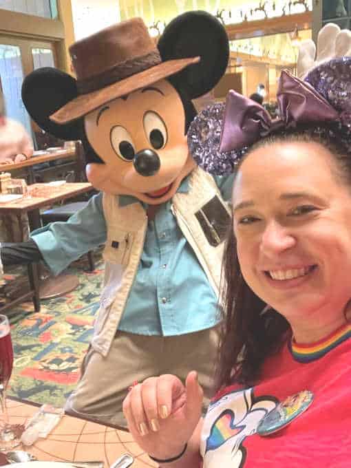 Woman in red shirt poses with Mickey Mouse who is wearing a camping outfit