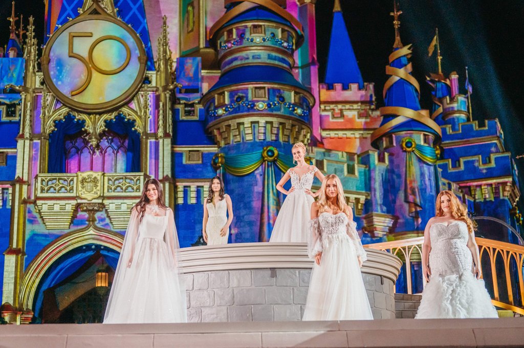 Five models wearing wedding gowns stand in front of Cinderella Castle