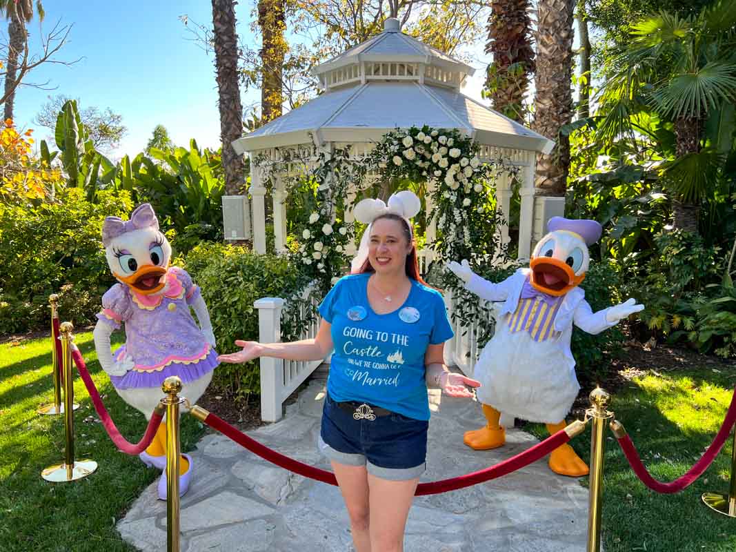 Woman in blue shirt and white Minnie Mouse ears poses with Daisy and Donald Duck at Adventure Lawn Gazebo