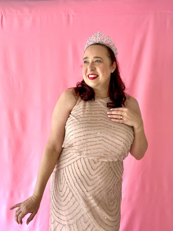 Woman wears beige form fitting gown with silver beading and a silver tiara