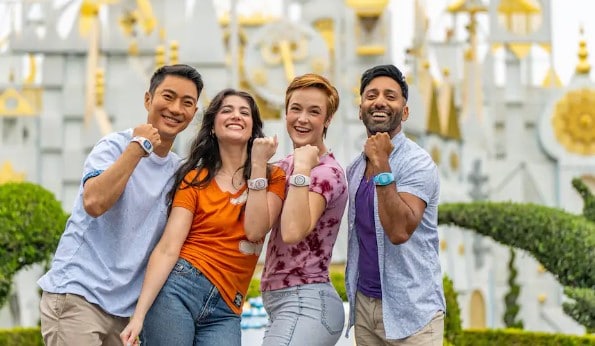 Four people standing in front of its a small world at Disneyland, holding up their arms to show they are wearing MagicBand+ bracelets