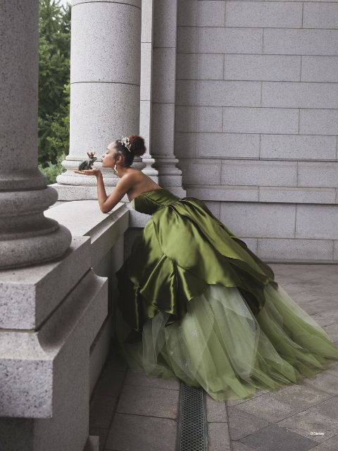 Woman wears dark and light green gown inspired by Princess Tiana; she is bending over and kissing a frog which she holds in her hands