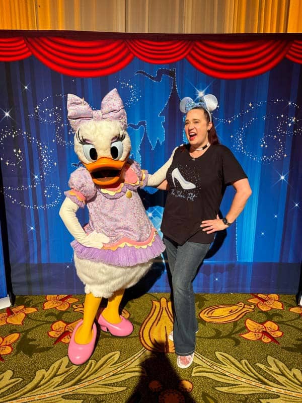 Woman in black shirt poses with Daisy Duck wearing a purple dress and hairbow in front of a blue backdrop