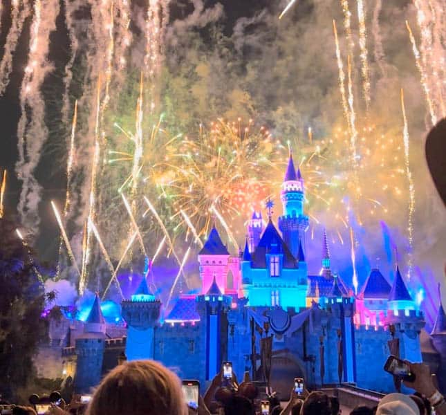 Sleeping Beauty Castle lit up with trans flag colors with fireworks in the sky at Disneyland Pride Nite