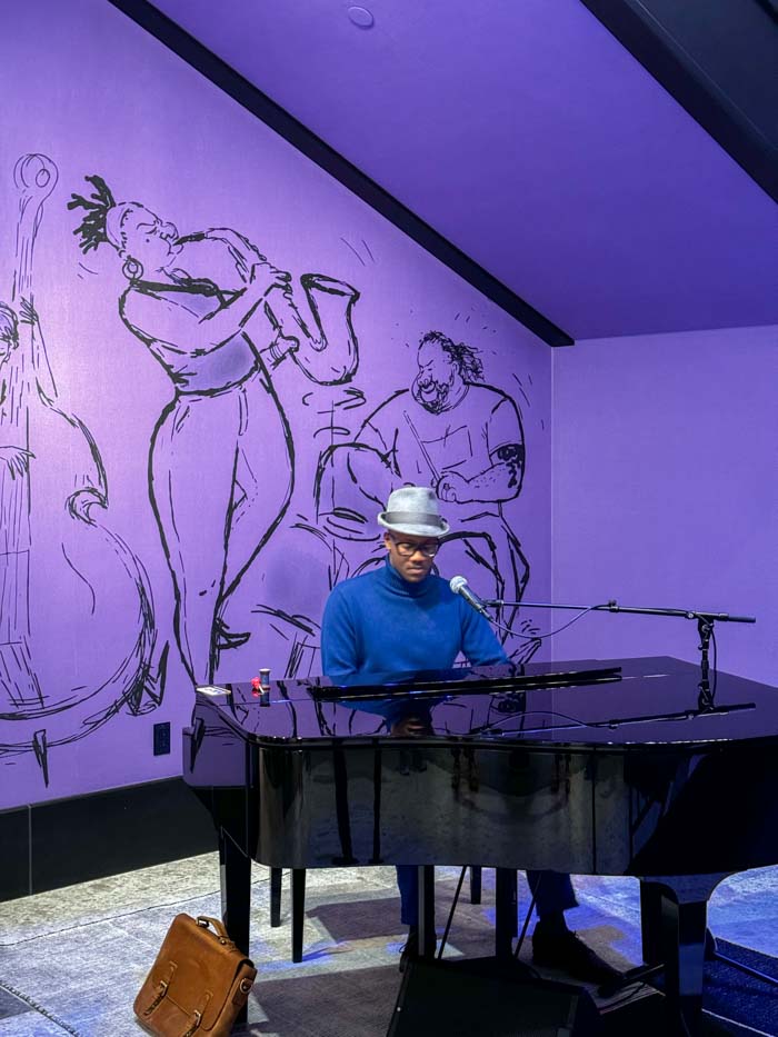 Joe Gardner plays a black piano in front of a purple mural. He is wearing a blue shirt and white hat.