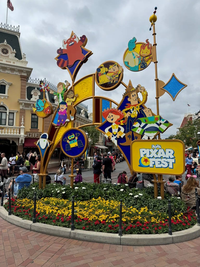 Colorful Pixar Fest sign featuring Pixar characters, surrounded by red and yellow flowers, with people and buildings in the background at Disneyland.