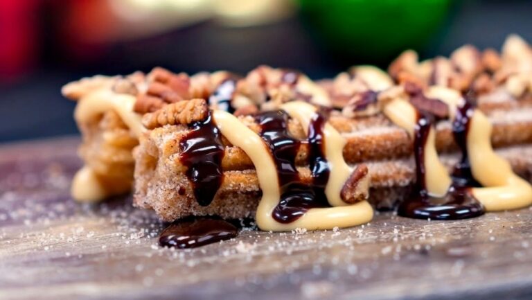 Close-up of Disneyland churros topped with chopped nuts, chocolate sauce, and a light-colored cream sauce, served on a wooden surface dusted with sugar.
